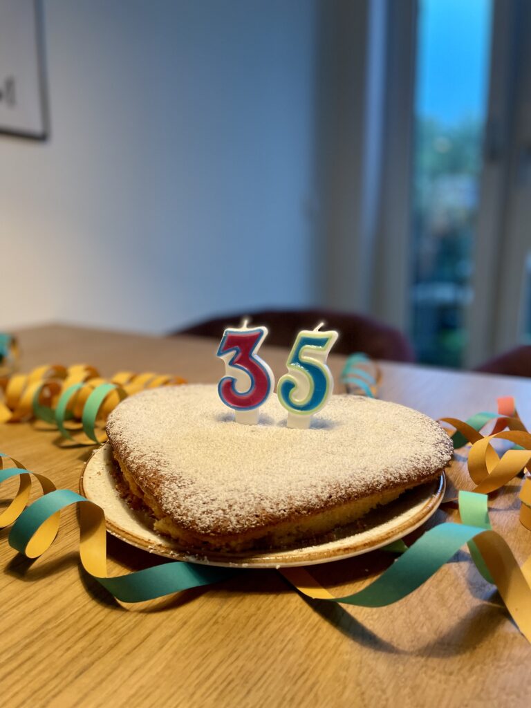 Herzförmiger Kuchen mit Puderzucker bestäubt, dekoriert mit bunten Geburtstagskerzen in Form der Zahl 35, auf einem Holztisch mit farbigen Luftschlangen.