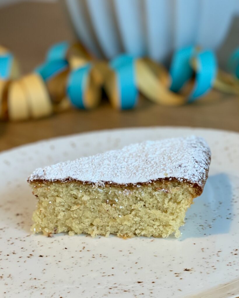 Ein Stück heller, saftiger mallorquinischer Mandelkuchen mit Puderzucker bestäubt auf einem weißen, gesprenkelten Keramikteller, im Hintergrund bunte Party-Luftschlangen.